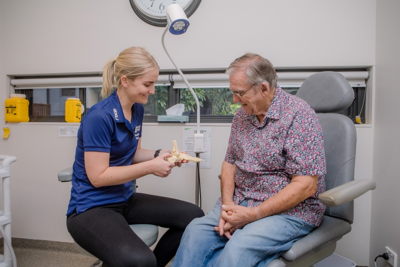 A senior man is sitting in a medical chair while a podiatrist is holding an anatomical foot model