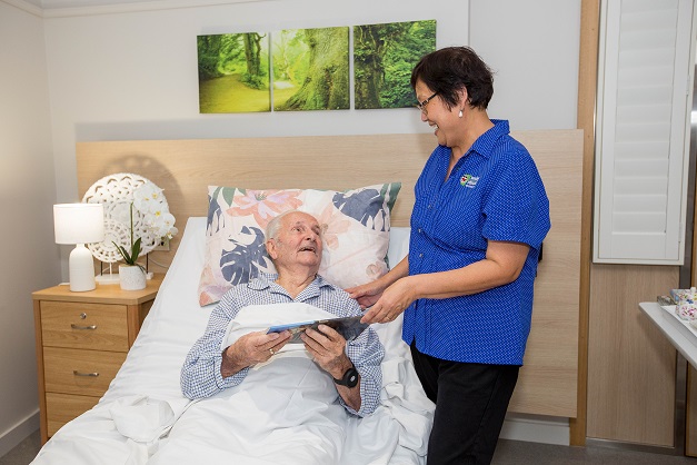 02082023_Respite_HaddenPlace_Promotional_018_resize A gentleman lying in bed and being handed a book by a carer