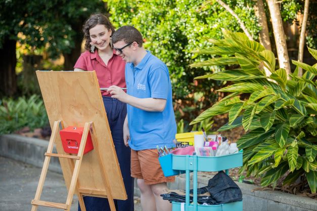 Young man painting outside on an art easel with his teacher