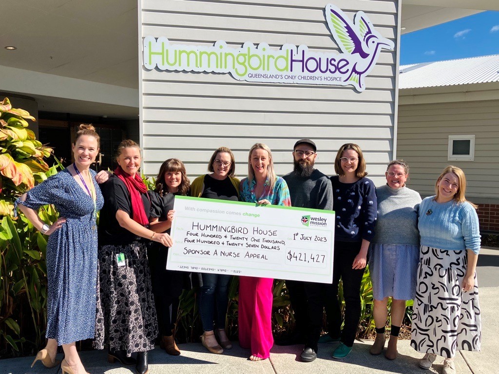 Eight women and a men stand with an oversized novelty cheque in front of a building with Hummingbird House signage