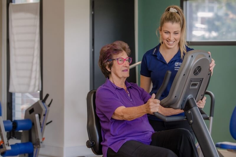 Woman assisting woman on exercise bike.